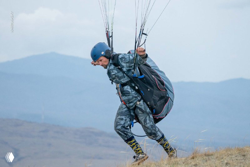 Пилот в каске и упаковке параплана делает разбег по склону перед взлётом; видны стропы и подготовка к старту на фоне горного ландшафта.