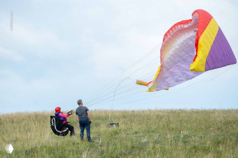 Инструктор рядом с учеником и надутым крылом яркой раскраски на травянистом склоне — сцена наземной подготовки и инструктажа перед полётом.