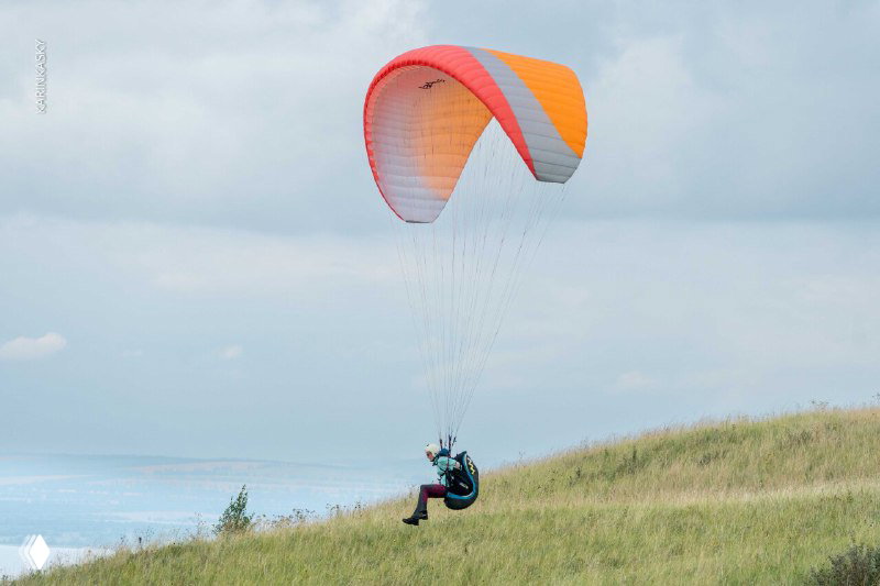 Параплан в полёте над пологим травянистым склоном: пилот висит в подвесной системе, крыло устойчиво держится в воздухе над холмом.
