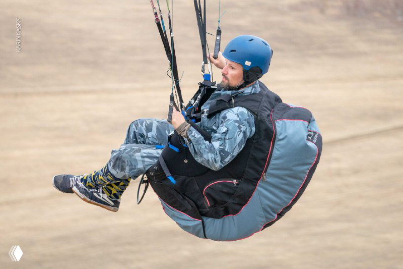 Крупный план пилота в подвесной системе и шлеме во время полёта: ручки управления, стропы и спокойный профиль пилота на фоне неба.