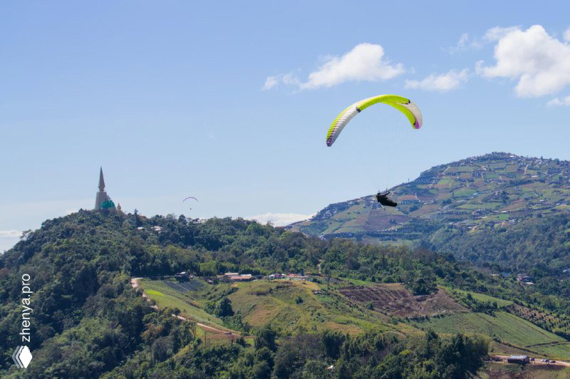 Два пилота на парапланах в небе на фоне холмов и храмовой башни, панорамный вид и мягкие облака в кадре.