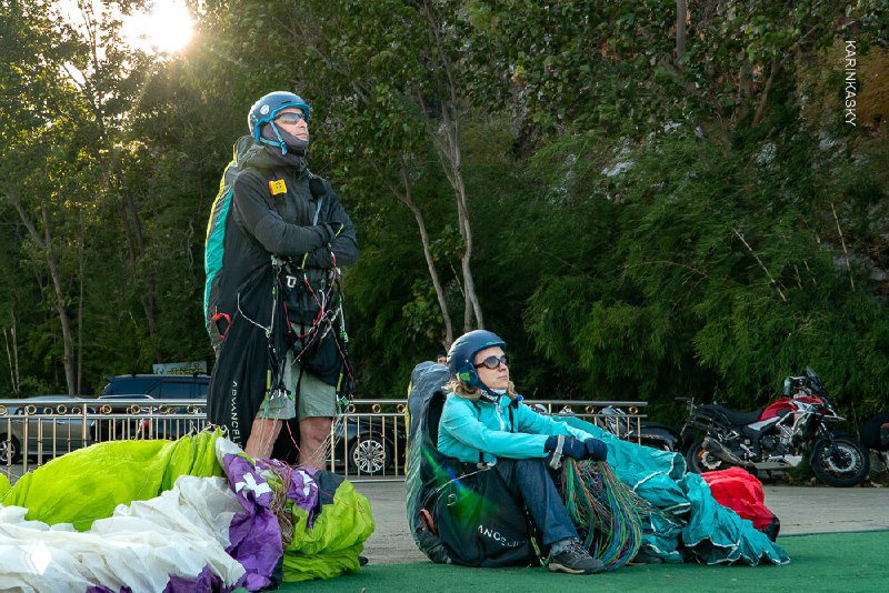 Два парапланерастa на старте, окружённые сложенным куполом и снаряжением — кадр подготовки с коллективной атмосферой.