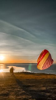 Параплан на склоне у озера Беле на закате: пилот готовит крыло к взлёту, видна береговая линия, спокойная вода и широкое небо