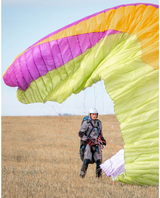 Пилот в каске и подвесной системе удерживает раскрытый яркий купол параплана на фоне степного поля во время наземной тренировки.