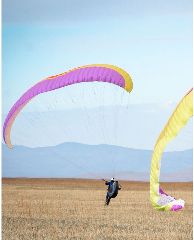 Несколько парапланов в воздухе над равниной Хакасии, виден стартующий пилот и линия отдалённых гор на горизонте в тренировочном полёте.