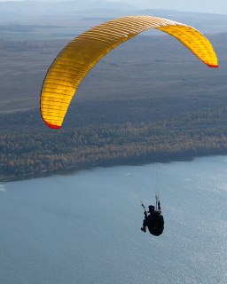 Параплан в полёте над лесистым берегом реки, силуэт пилота в подвесной системе на фоне осеннего пейзажа — учебный полёт в Хакасии.