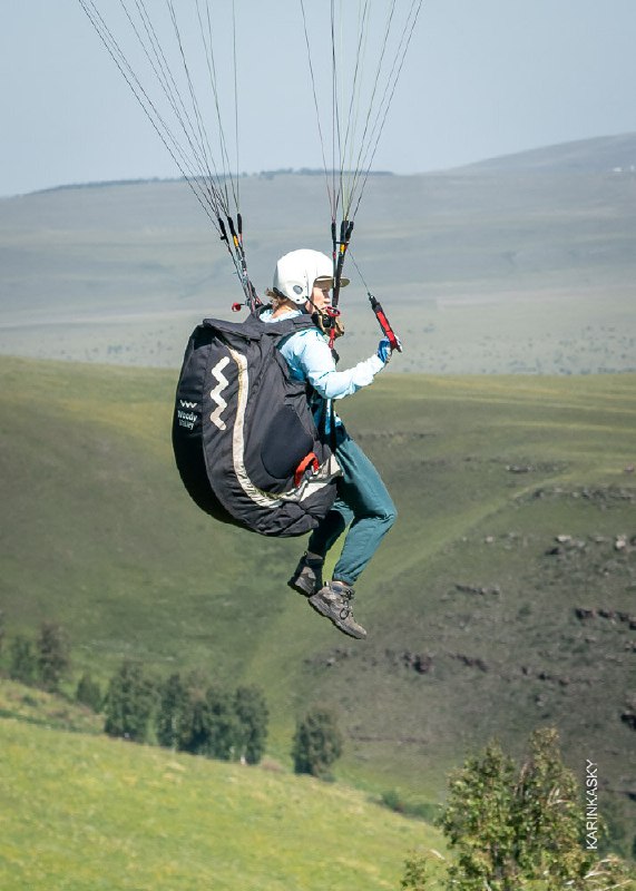Пилот в шлеме висит над холмом на подвесной системе параплана; видно рюкзак-парашют и просторный пейзаж, подчёркивающий условия полёта.