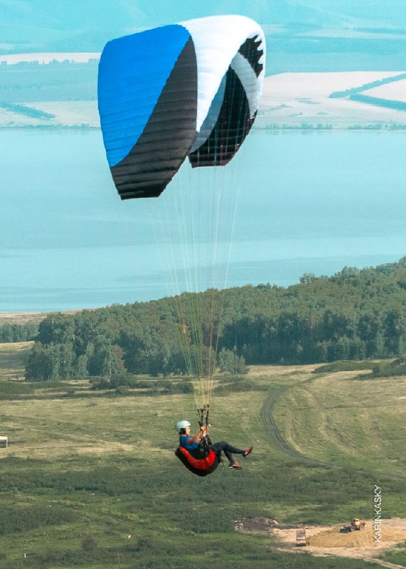 Два пилота в тандеме на парапланах над озером и лесом: раскрытые крылья, видна водная поверхность и береговая полоса вдалеке.