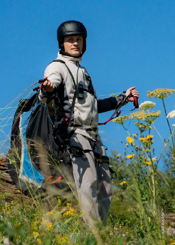 Пилот в защитном шлеме стоит на цветущем склоне, держит стропы параплана; на заднем плане видно летное поле и летнюю растительность.
