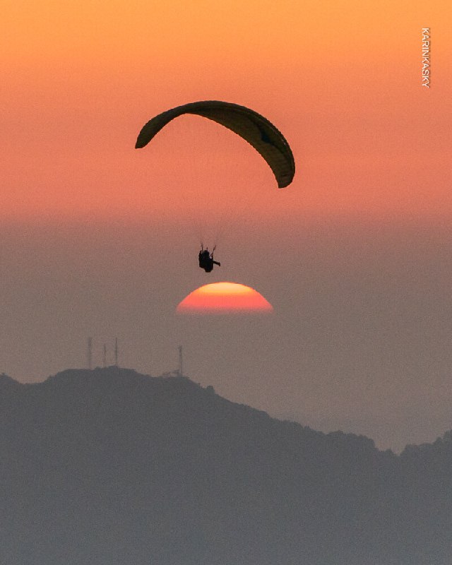 Один парапланист на фоне заходящего солнца и оранжевого неба; силуэт на фоне горного горизонта в момент заката.