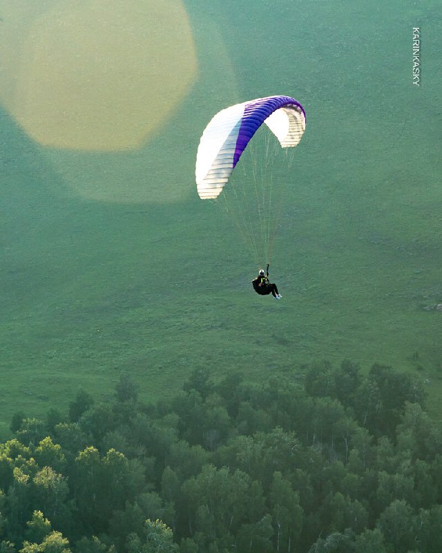 Один парапланист в полёте над зелёной равниной; вид сверху показывает склон и поля внизу, спокойные условия для тренировки.