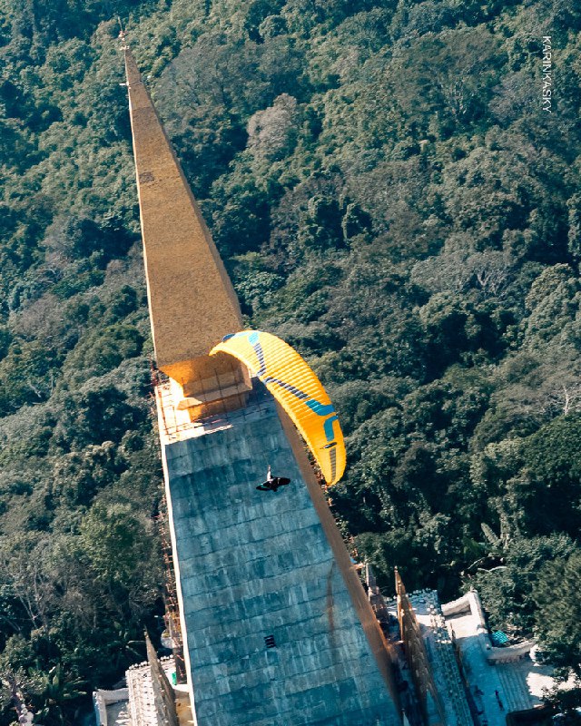 Парапланист с жёлтым крылом выполняет пролёт около бетонной конструкции или дамбы — вид близкого пролёта вдоль архитектурного объекта.