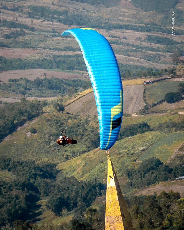 Ярко-голубой параплан в полёте над сельской местностью; вид сверху показывает поля, деревья и рекреационные зоны под крылом.