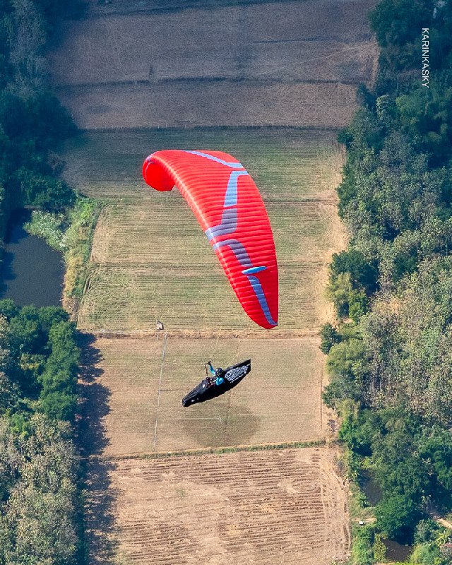 Красный параплан в ровном полёте над пашней; композиция подчёркивает форму крыла и пилота на фоне земляного рельефа.