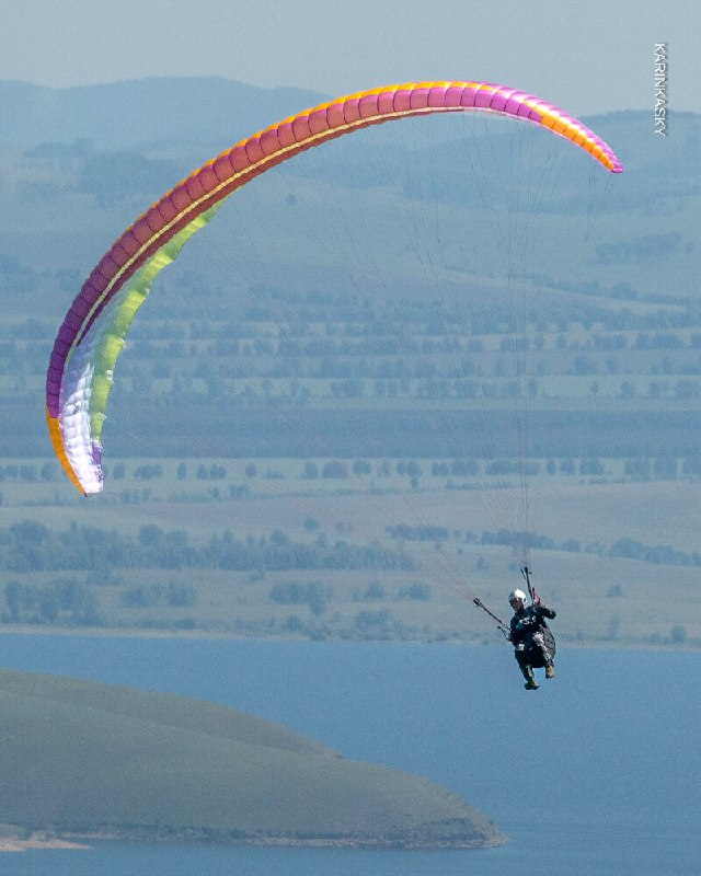 Одинокий пилот под разноцветным куполом на фоне дальних холмов и водоёма, изображение подчёркивает высоту и маршрутный полёт в термике.