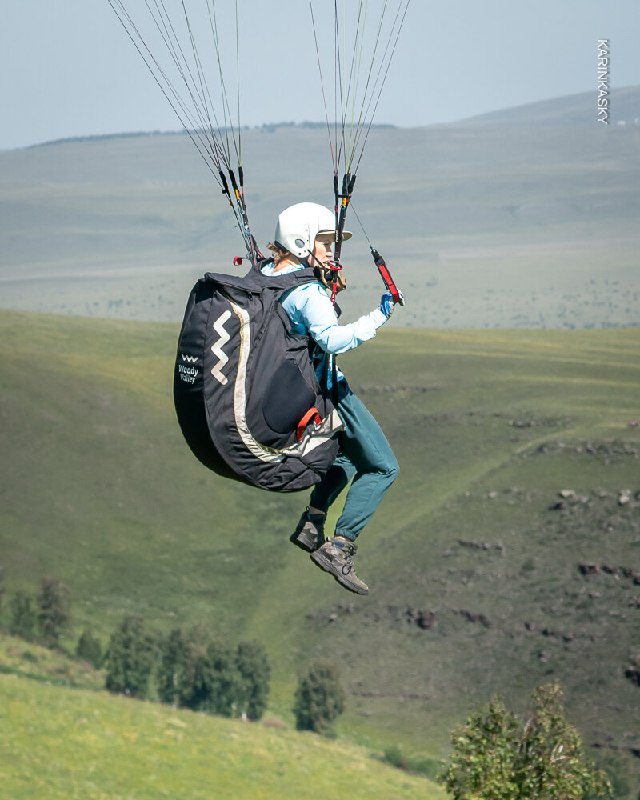 Пилот в шлеме и подвеске на взлёте или наборе высоты над холмом; вид сбоку показывает подвесную систему и технику запуска или контролируемого набора.