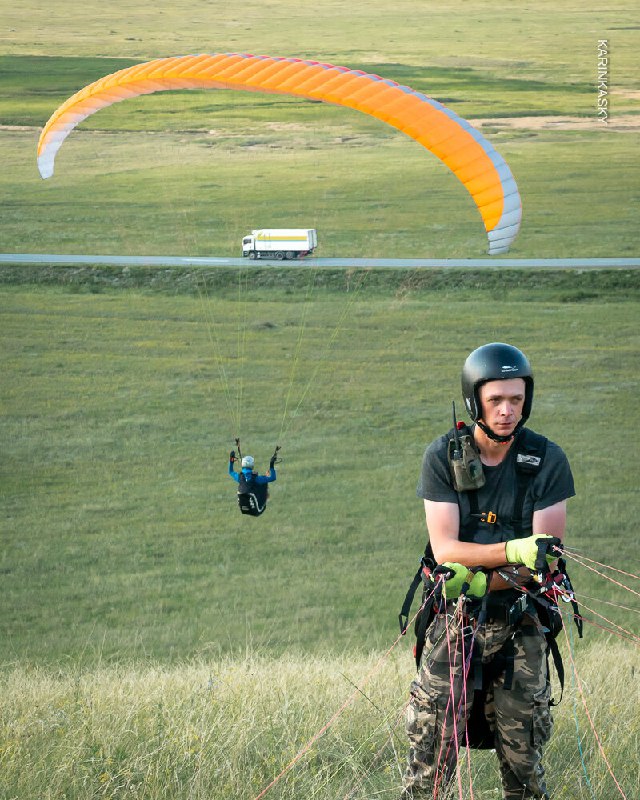 Оранжево‑жёлтый параплан в полёте над трассой и автобусом вдоль поля; композиция показывает масштаб и близость к земле при среднем полёте.