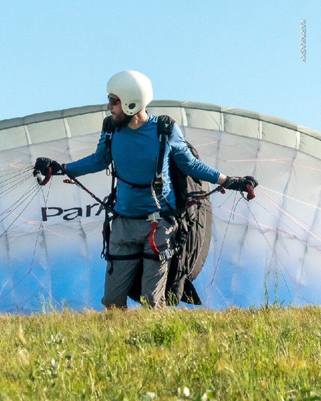 Инструктор или пилот на старте рядом со сложенным куполом на земле; кадр подчёркивает подготовку экипировки перед взлётом на травянистом склоне.