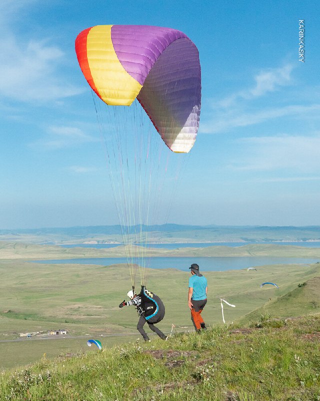 Пилот в голубой футболке бежит при старте, за ним видно несколько крыльев и открытые ландшафты — момент взлёта с журнала тренировки в степи.
