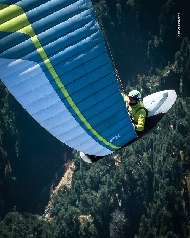 Пилот в зелёной экипировке под сине‑зелёным крылом, полёт на фоне лесистых склонов и неба; кадр подчёркивает погодные условия и рельеф.