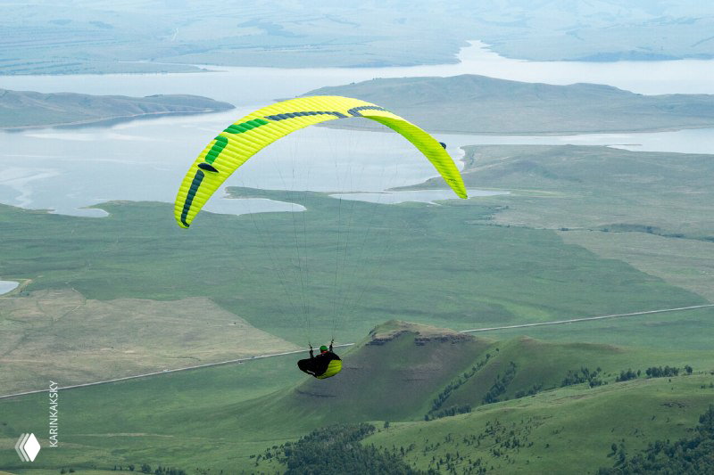 Зелёный параплан над широкими заливными лугами и извилистым озером — открытый степной рельеф, возможный маршрут в Хакасии с хорошей видимостью и пространством для манёвра.