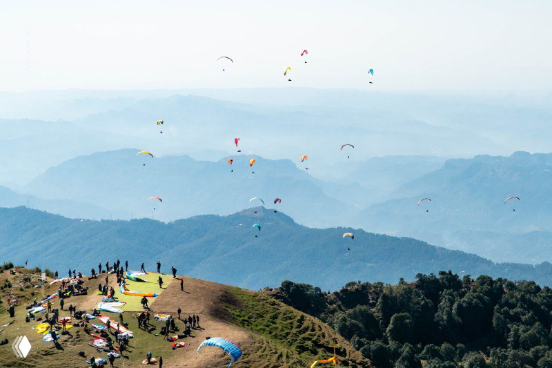 Много парапланов в небе над горным хребтом; на переднем плане стартовая площадка с участниками, парашютами и подготовкой к массовому вылету.