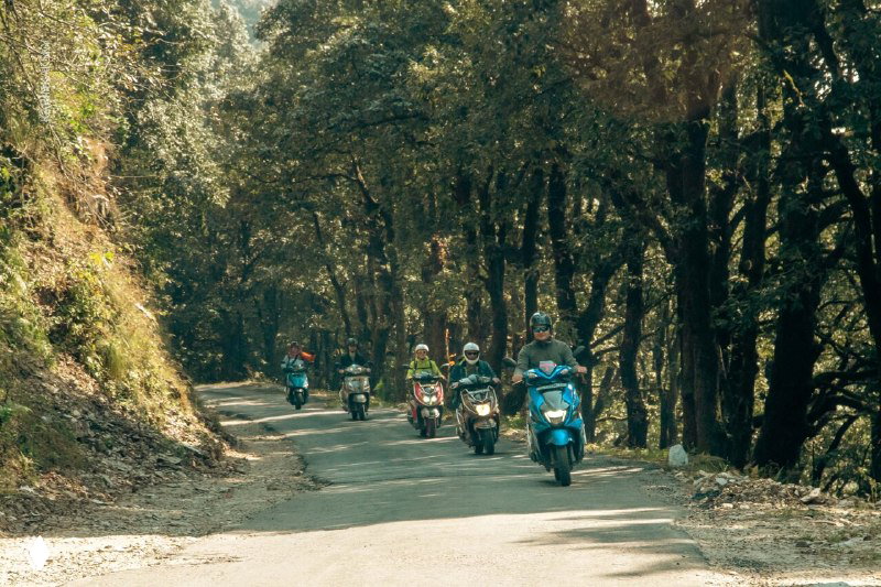 Горная дорога сквозь лес с колонной мотоциклистов и скутеров; транспортная логистика и подъёмы к точкам старта в горном рельефе.