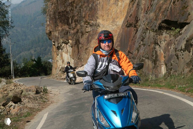 Пилот в экипировке на синем скутере у скалистой горной дороги; сцена подготовки и передвижения по маршруту полётов в горах.