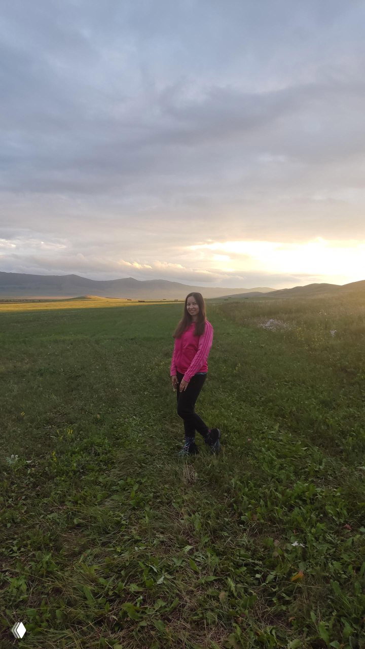 Женщина в розовой куртке стоит на зелёном поле в Хакасии при закате, на фоне низких холмов и рассеянных облаков над равниной.