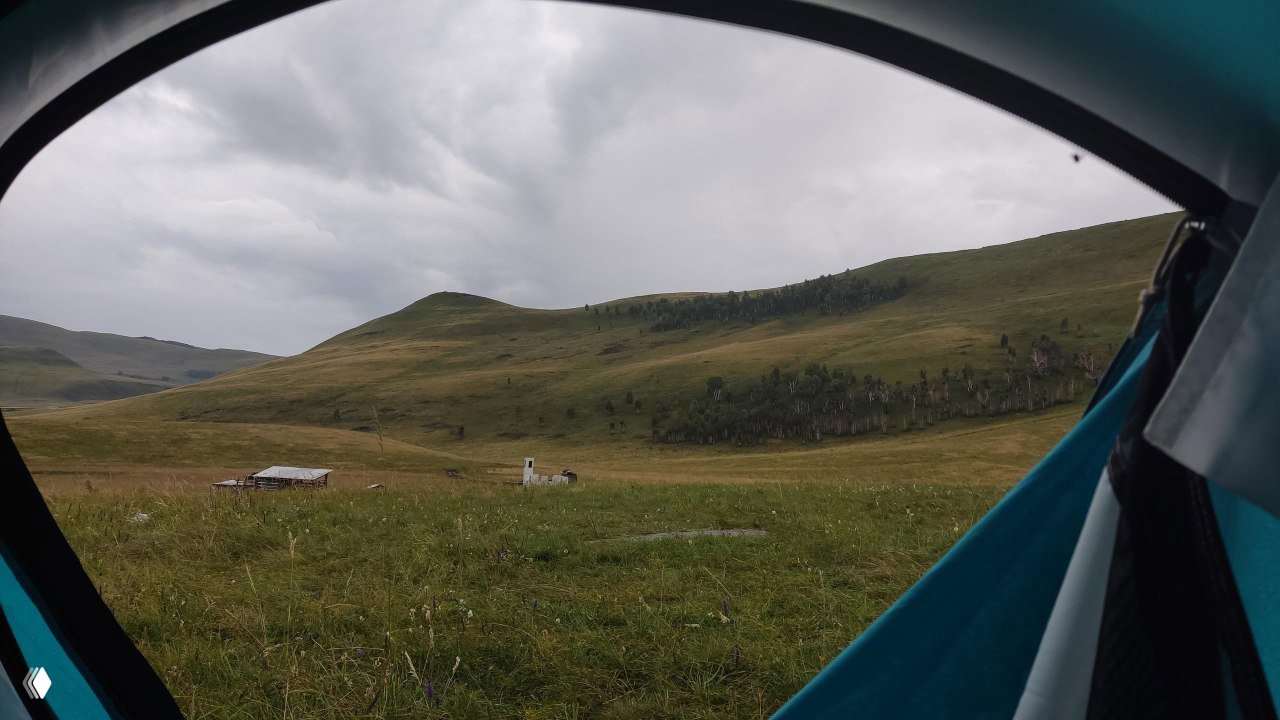 Вид из палатки на широкие зелёные луга и пологие холмы Хакасии, пасмурное небо и несколько хозяйственных построек вдали.