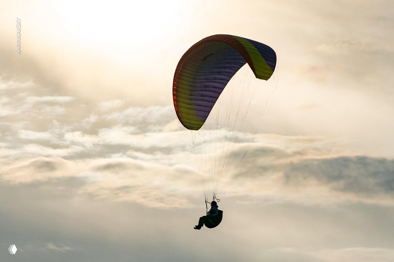 Парапланист в полёте на фоне светлого неба: силуэт пилота под ярким куполом параплана в профиль, момент парения над ландшафтом.