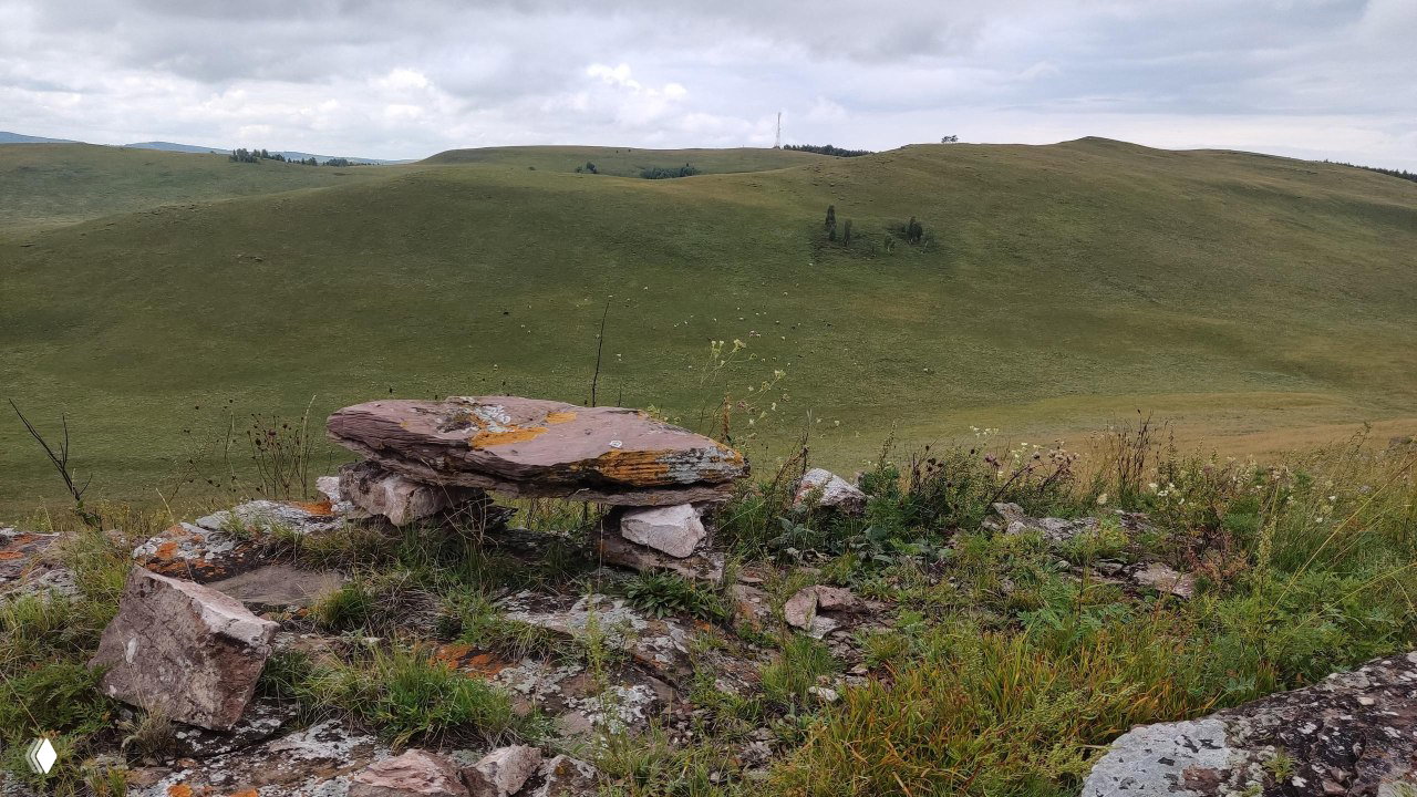 Каменные плиты на вершине холма и травянистый склон в Хакасии, вид на удалённые пастбища и плавные контуры холмов под облаками.