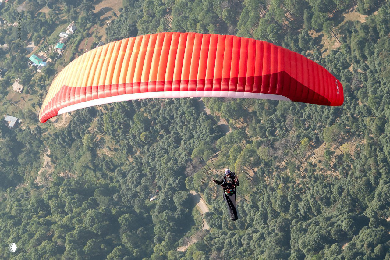 Пилот на красном параплане в воздухе над лесистой местностью, вид сверху подчёркивает простор и аэродинамику крыла во время полёта