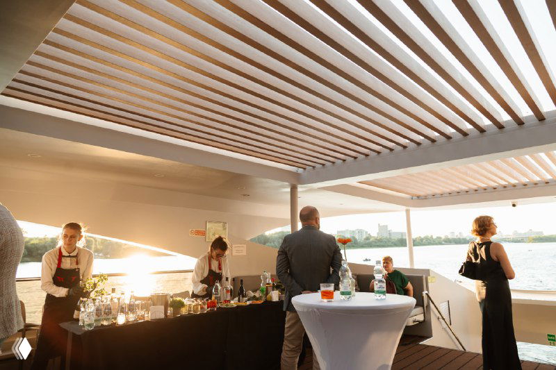 Бар и зона обслуживания на палубе яхты во время презентации, официанты и гости на фоне реки при закате, атмосфера приёма.