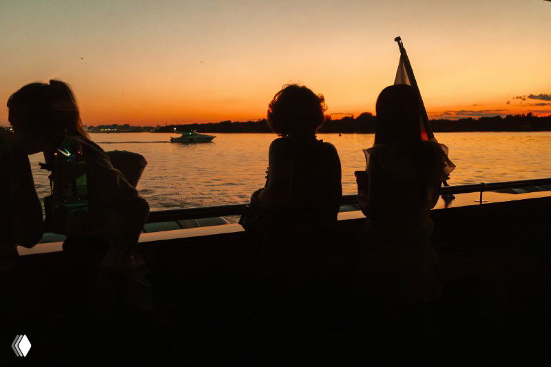 Силуэты гостей на фоне заката над рекой: съемка с палубы яхты подчёркивает вечернюю атмосферу презентации жилого комплекса.