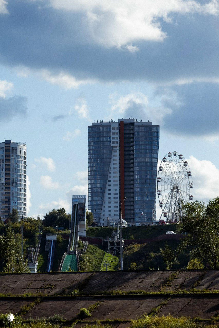 Городской пейзаж с высоты — жилой дом и колесо обозрения на фоне зелёных склонов: вид, демонстрирующий окружение ЖК и близкую инфраструктуру.