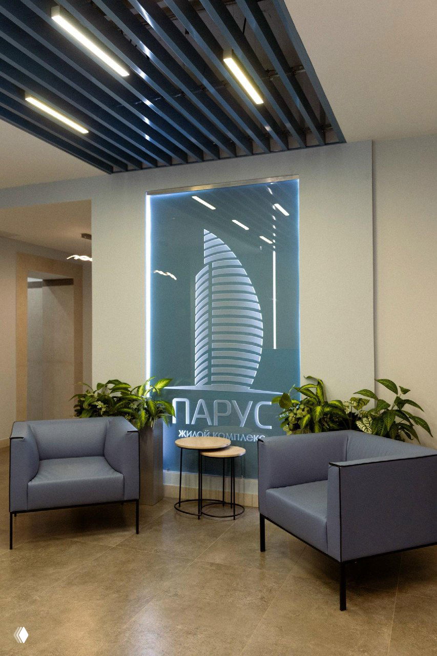 Ресепшн и интерьер демонстрационной зоны ЖК «Парус»: логотип комплекса в световой панели, кресла и зелёные растения в лобби.