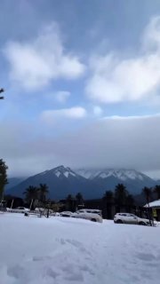 Вертикальный видеокадр: заснеженная парковка и припаркованные машины на фоне гор с низкими облаками — пейзаж рядом с жильём.