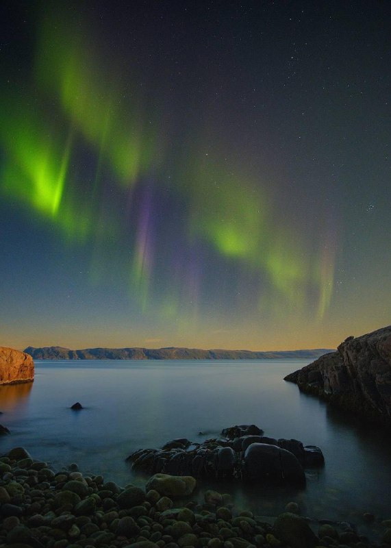 Полярное сияние над каменистым побережьем ночью: зелёные и фиолетовые полосы света над спокойной водой — мистическая сцена вдохновения.