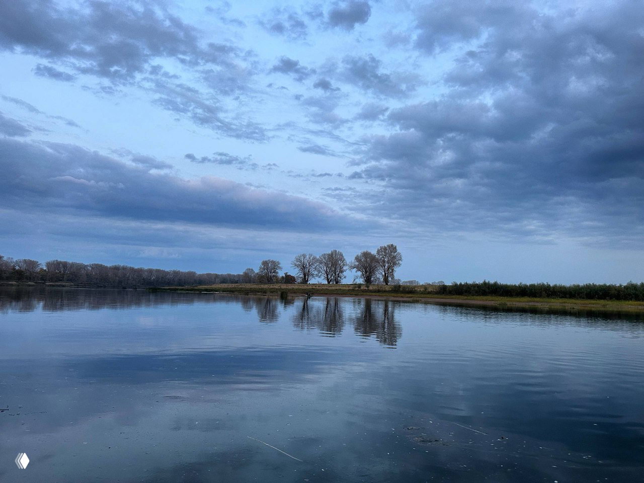 Кадр озера с прибрежными зарослями и гладкой поверхностью воды; небо покрыто плотными облачными слоями, придающими сцене драматичный тон.