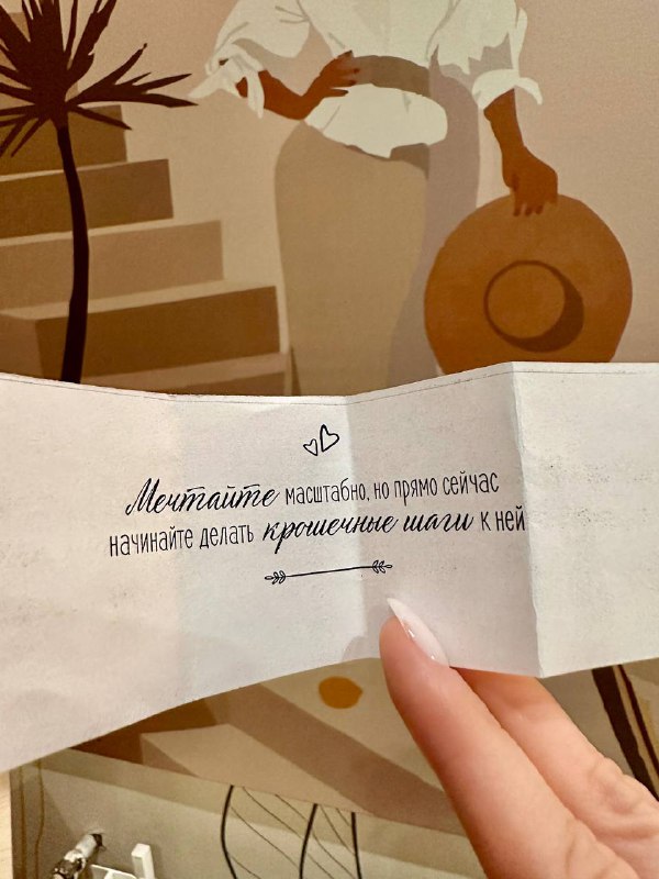 Рука держит бумажку с предсказанием на фоне настенной росписи и элементов декора в кафе — добрый завершающий штрих к визиту.