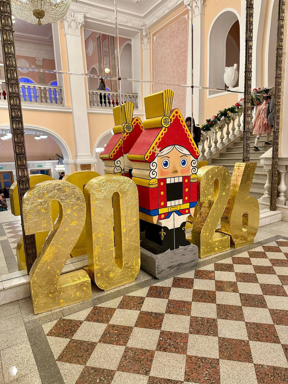 Декоративные объёмные цифры «2026» и деревянный щелкунчик у зеркальной стены в фойе Театра Оперы и Балета Уфа, ступени и мраморный пол рядом.