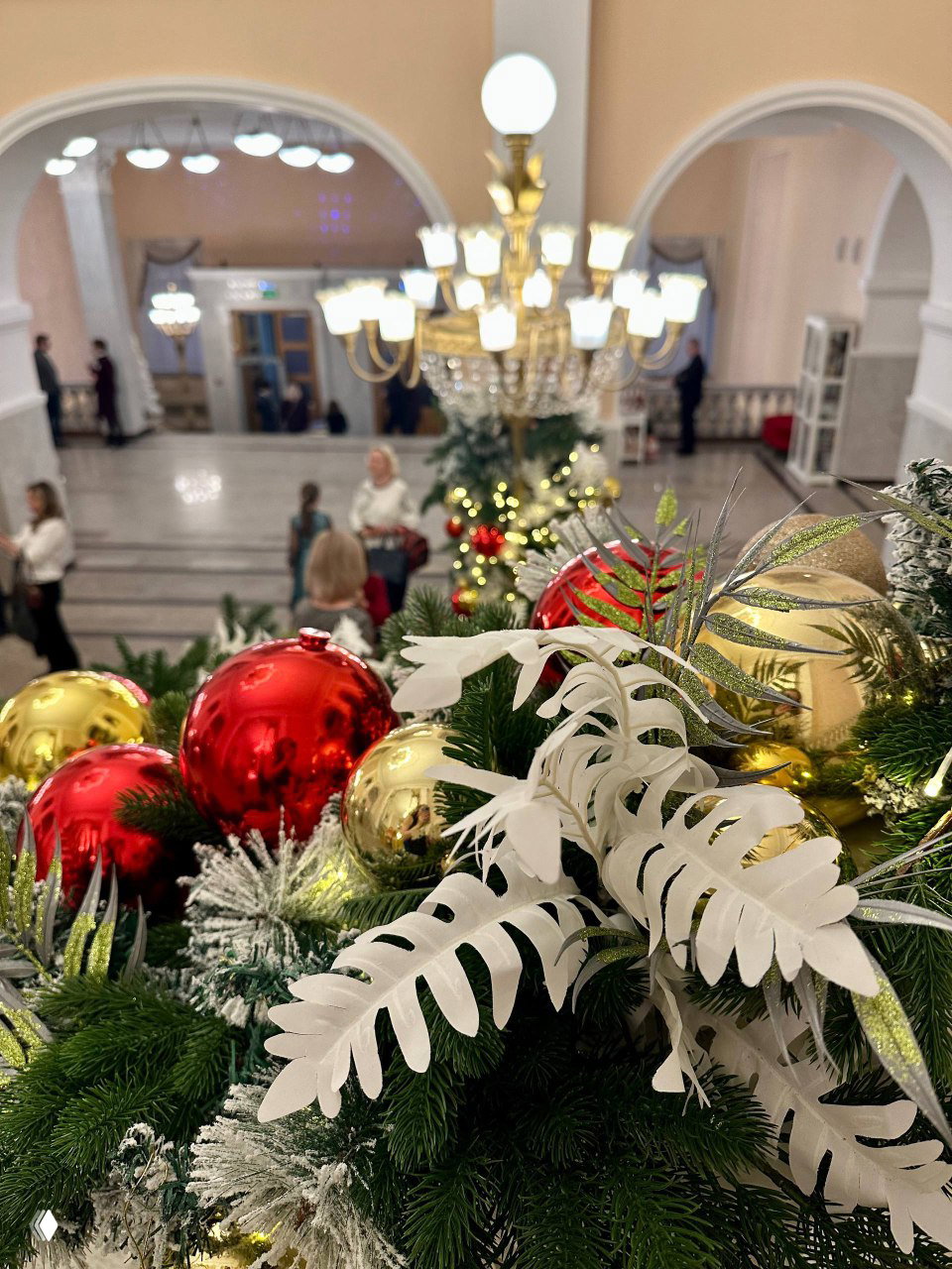 Крупный план праздничной гирлянды с красными и золотыми ёлочными шарами и искусственной зеленью на фоне размытых посетителей и люстры фойе.