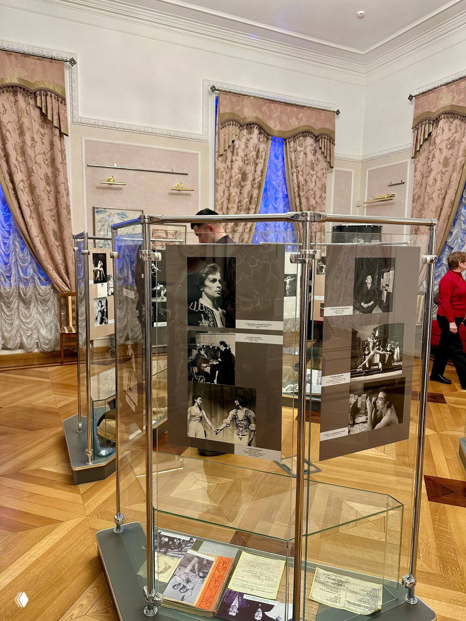 Стеклянные выставочные стенды с чёрно‑белыми фотографиями и документами о карьере Рудольфа Нуриева в зале театра с паркетом и портьерами.