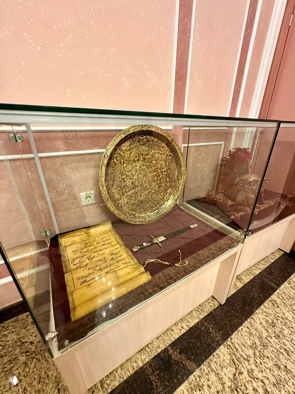 Витрина с декоративным щитом, свитком и коротким мечом: старинные мотивы и филигранная роспись на предметах реквизита в музее.
