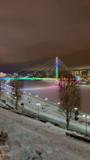 Ночная панорама набережной реки Тура в Тюмени: освещённый Мост Влюблённых, снег на берегу, огни и отражения в воде.