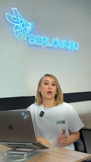 Женщина-лектор за столом с ноутбуком на фоне неоновой вывески «BERLOVA.RU», кадр из видеопрезентации о маркетинге клиник.