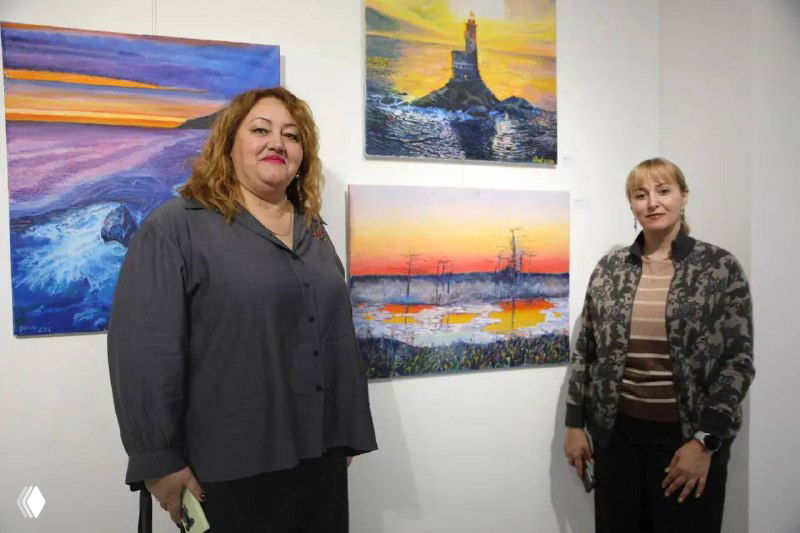 Две женщины на фоне выставочных полотен Игоря Буреева в белом зале галереи: одна держит документы, вторая стоит рядом, позируя у работ.