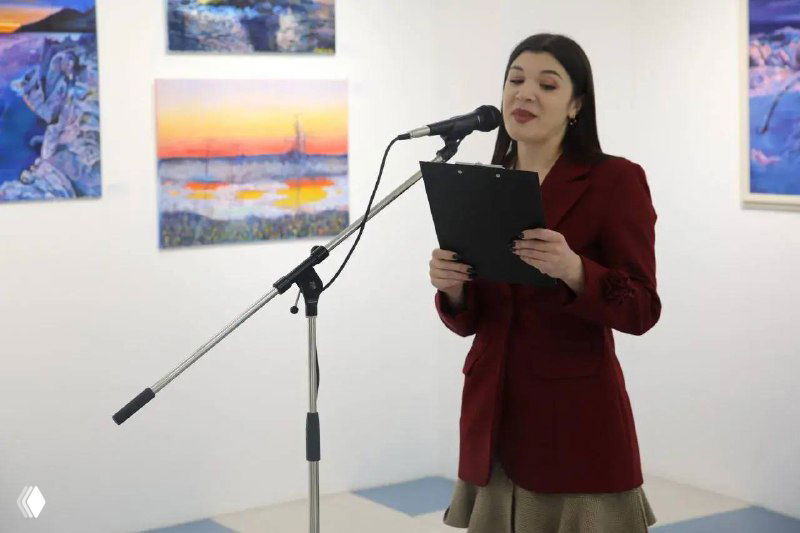 Женщина выступает у микрофона на открытии выставки Игоря Буреева; на заднем плане видны картины с маяком и закатом, сцена вернисажа в галерее.