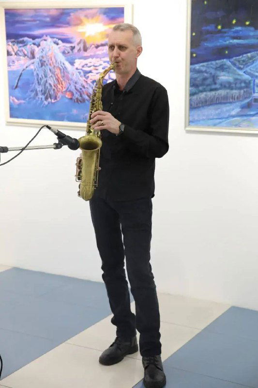 Музыкант с саксофоном исполняет живую музыку на вернисаже выставки в галерее «Город художников», посетители слушают в белом пространстве музея книги Чехова.